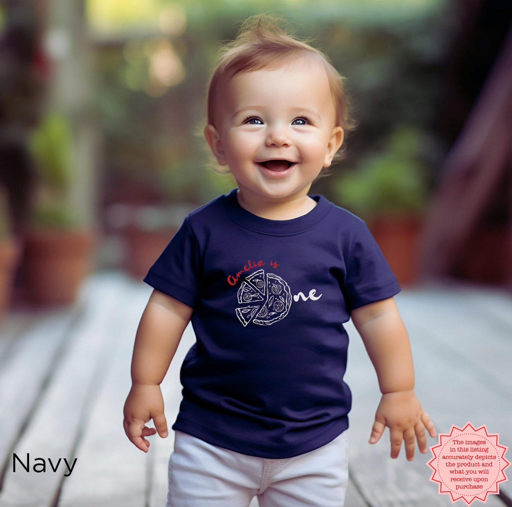 Happy toddler wearing navy blue personalized pizza party t-shirt with ‘Amelia is One’ graphic, custom Italian first birthday outfit for babies and toddlers.