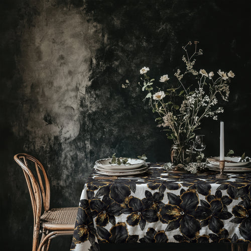 Modern botanical black orchid tablecloth draped over a rectangular dining table, featuring painterly black florals with subtle gold detailing on an ivory background