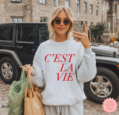 Woman wearing ash grey C’EST LA VIE sweatshirt, French quote crewneck, cozy Paris-inspired travel outfit