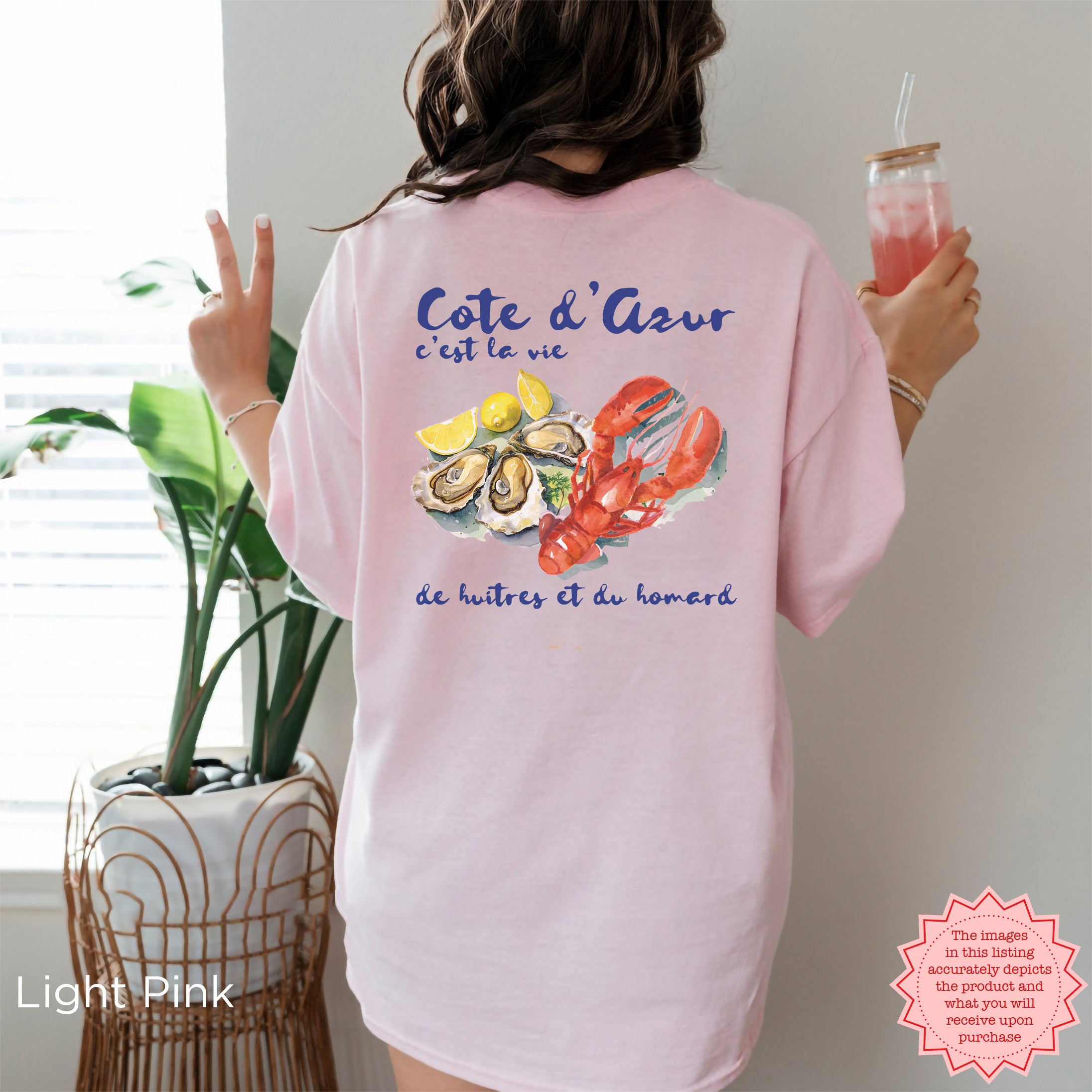Light pink Cote d’Azur lobster and oysters graphic tee, casual summer beachwear, featuring watercolor coastal design, worn indoors holding a pink drink, perfect lobster lover gift.