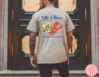 Sand and white Cote d’Azur coastal graphic t-shirt featuring watercolor lobster and oysters, worn by two friends at the beach enjoying pizza, ideal for summer vacation and seafood lover style.