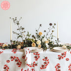 Mediterranean-inspired Tomato Block Print Tablecloth on a dining table with neutral plates and a vase of flowers. The vibrant tomato design adds a colorful and fresh touch to the table setting.