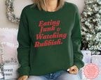 Christmas sweatshirt styled with snacks and a cosy movie-night setting, featuring humorous retro holiday lettering.