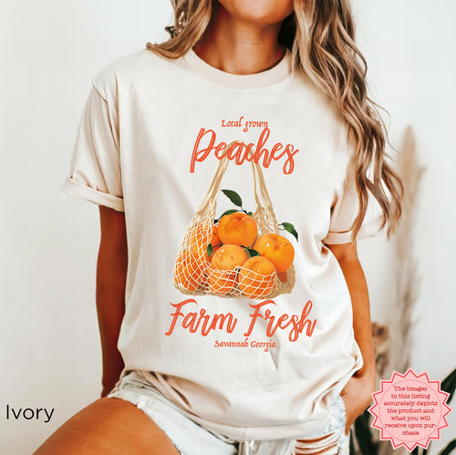 Model wearing a vintage-inspired Farm Fresh Peaches graphic T-shirt featuring a market produce bag filled with peaches, leafy green details, and “Local Grown,” “Farm Fresh,” and “Savannah Georgia” lettering.