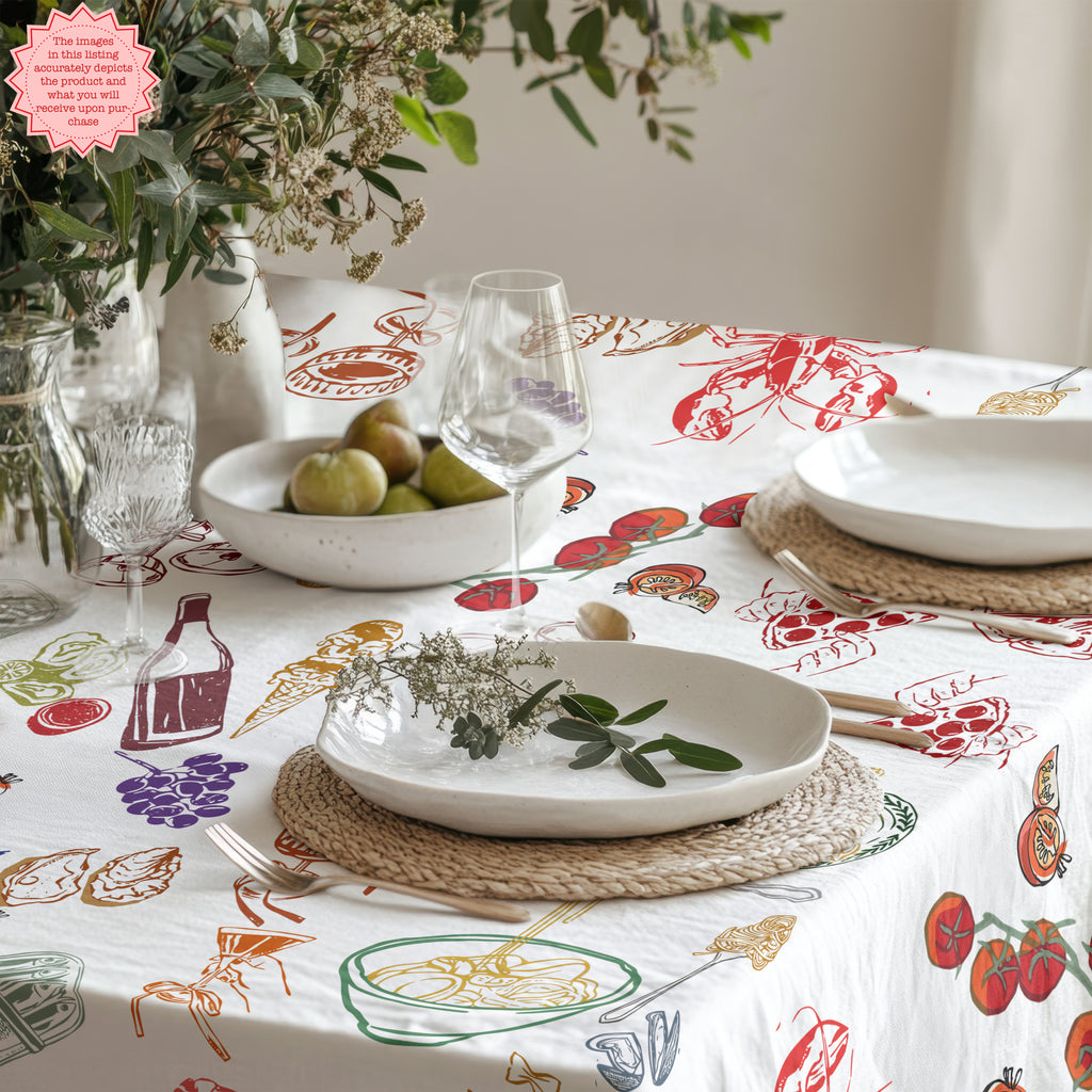 Modern Mediterranean dining table styled with a whimsical Italian summer tablecloth featuring illustrated food designs, ceramic dinnerware, fresh flowers and European-inspired décor.