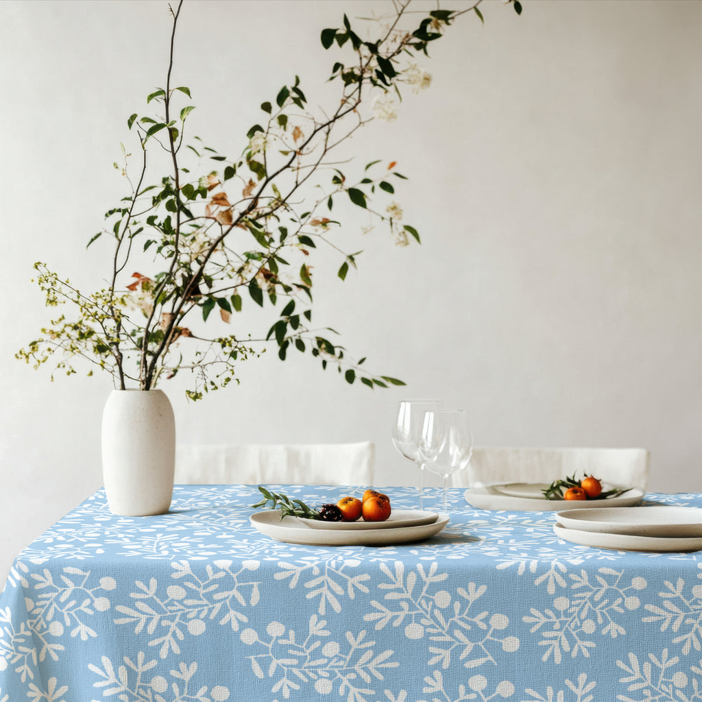 Mediterranean-inspired olive branch tablecloth in light blue on dining table with greenery, ceramic plates, and wine glasses.