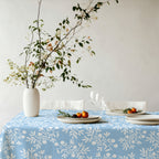 Mediterranean-inspired olive branch tablecloth in light blue on dining table with greenery, ceramic plates, and wine glasses.