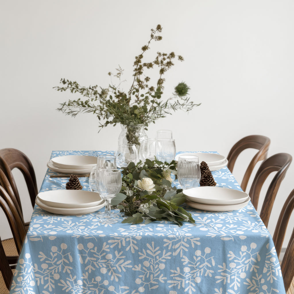 Mediterranean-style dining table with light blue olive branch tablecloth, neutral plates, and a vase of white flowers.