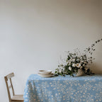 Elegant Mediterranean table setting with olive branch tablecloth in light blue, adorned with white roses and simple ceramic plates.
