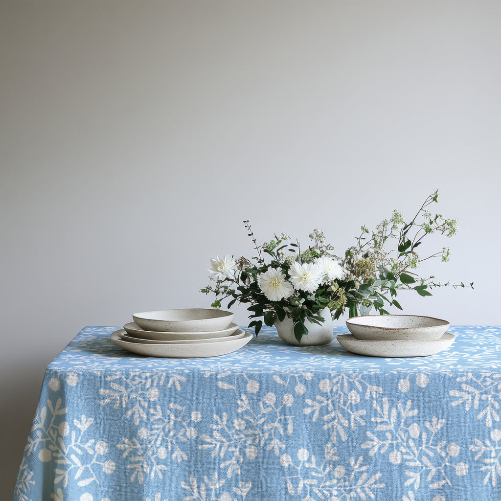 Casual Mediterranean dining table with light blue olive branch tablecloth, set with ceramic plates and fresh flowers for a natural look.