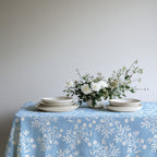 Casual Mediterranean dining table with light blue olive branch tablecloth, set with ceramic plates and fresh flowers for a natural look.