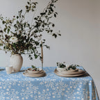 Modern Mediterranean dining table with olive branch tablecloth in light blue, set with wine glasses, rustic plates, and fresh flowers.