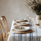 French country inspired dining setting with a heritage blue stripe tablecloth, neutral ceramics, woven textures, and relaxed Mediterranean styling.