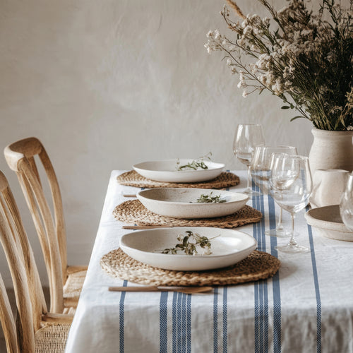 French country inspired dining setting with a heritage blue stripe tablecloth, neutral ceramics, woven textures, and relaxed Mediterranean styling.