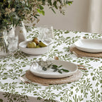Mediterranean olive branch tablecloth styled with white dinnerware, glassware, and a lush greenery centerpiece in a modern dining room.