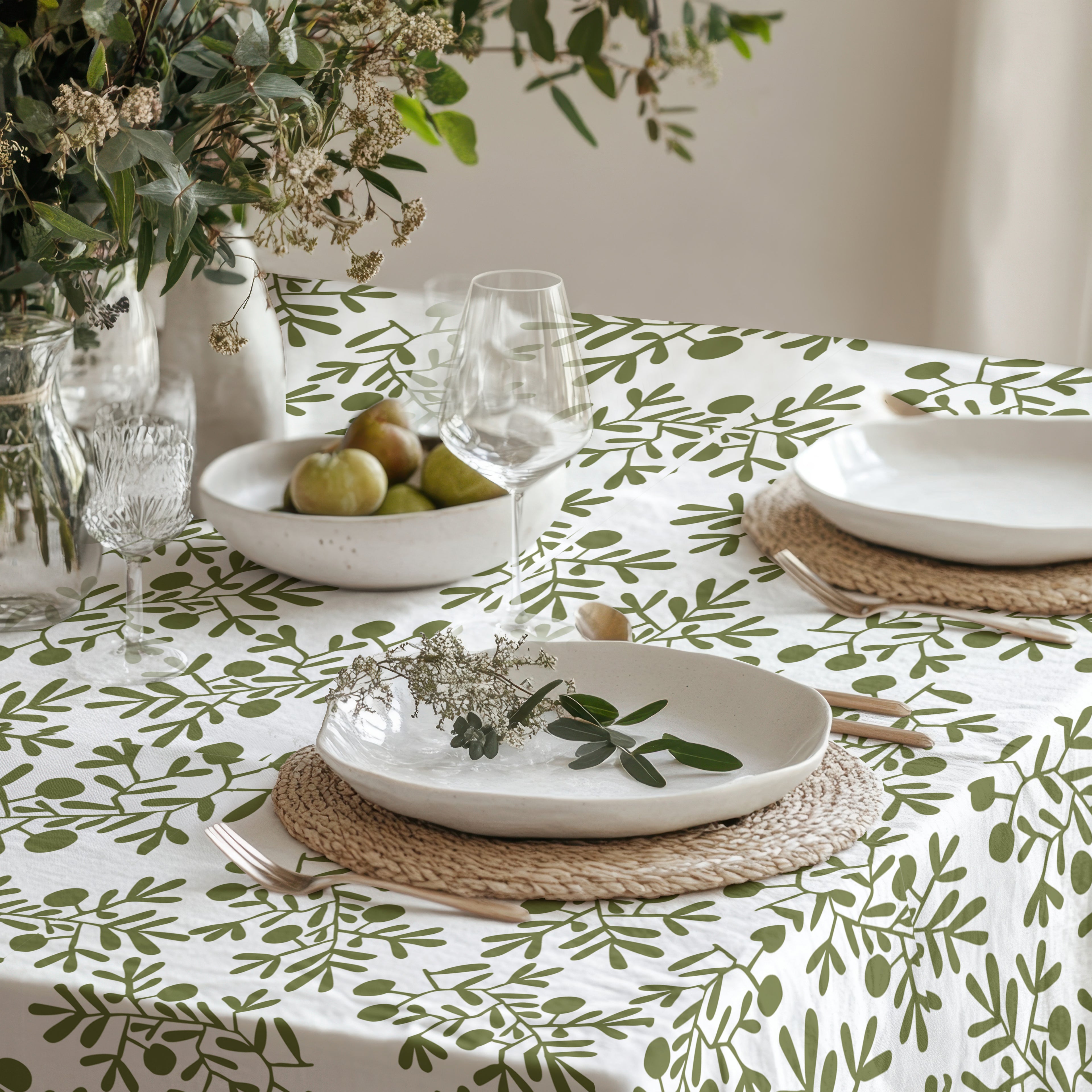 Mediterranean olive branch tablecloth styled with white dinnerware, glassware, and a lush greenery centerpiece in a modern dining room.