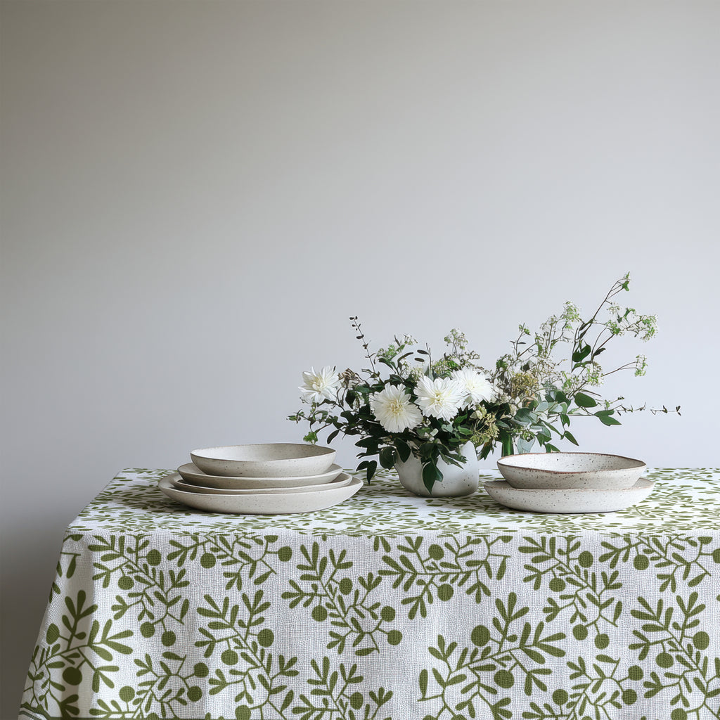 Modern Mediterranean dining table styled with an olive branch tablecloth, woven placemats, ceramic plates, and natural floral arrangements.