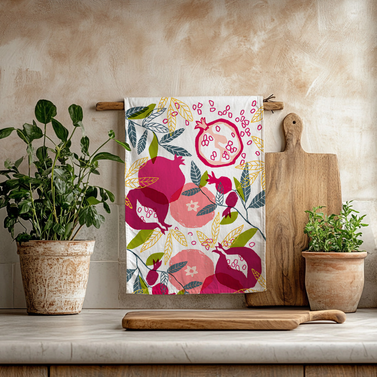 Painted pomegranate tea towel hanging in a bright Mediterranean kitchen with wooden utensils and greenery
