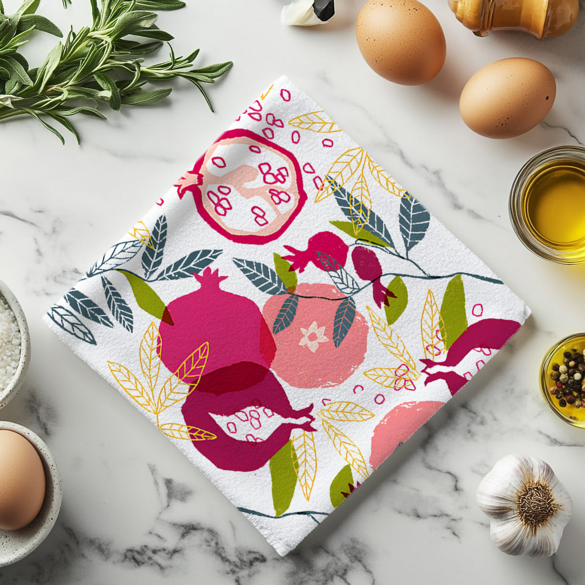 Colorful pomegranate tea towel with painted fruit design styled on marble countertop with eggs and olive oil