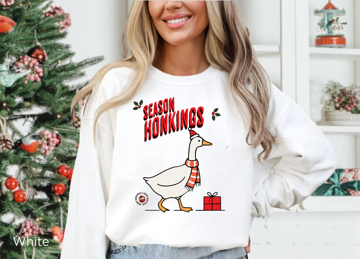 Woman wearing a White Season Honkings Christmas sweatshirt with a festive goose graphic while enjoying a coffee outdoors.