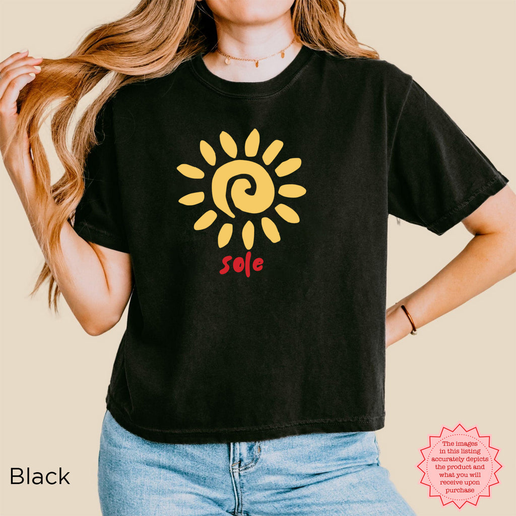 Woman wearing a black women's boxy graphic T-shirt featuring a golden sun illustration and hand-lettered “sole” design, a minimalist Mediterranean-inspired summer look.