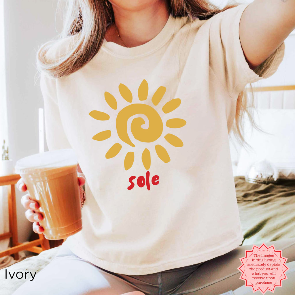 Woman wearing a cream boxy graphic T-shirt featuring a golden sun illustration and hand-lettered “sole” design.