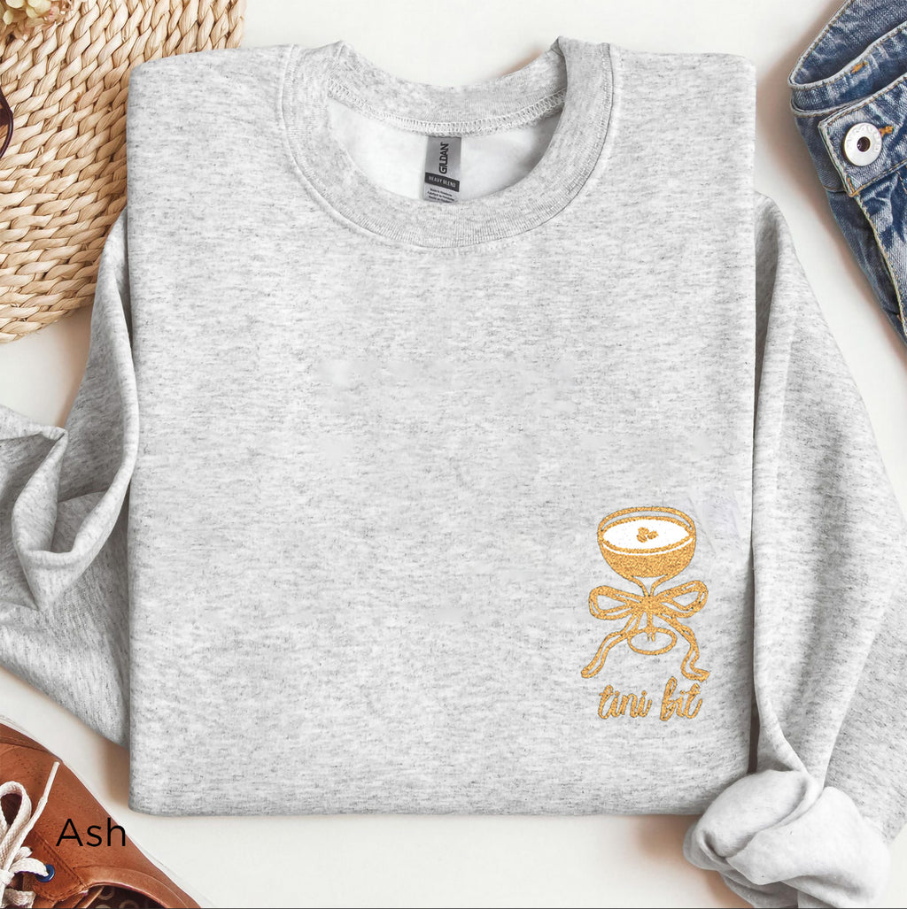 Folded “A Tini Bit Older” embroidered martini sweatshirt in ash, shown with rolled sleeves and paired with casual brown shoes, highlighting cozy birthday outfit for women.