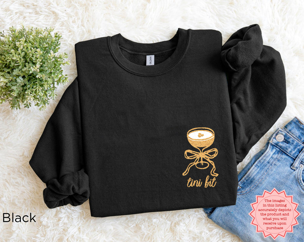 Folded black “A Tini Bit Older” embroidered martini sweatshirt on white textured surface with jeans and greenery, cozy bachelorette party sweatshirt for women.