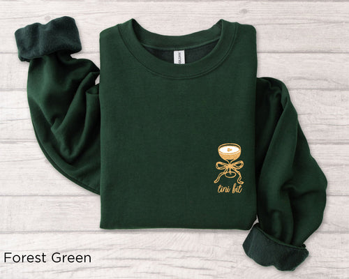 Folded forest green “A Tini Bit Older” embroidered martini sweatshirt, rolled sleeves, styled with cotton stems and wooden props, perfect women’s bachelorette or birthday gift.