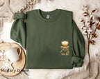 Folded military green “A Tini Bit Older” embroidered martini sweatshirt, rolled sleeves, displayed with neutral decor, casual cozy outfit for cocktail or birthday celebration.