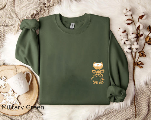 Folded military green “A Tini Bit Older” embroidered martini sweatshirt, rolled sleeves, displayed with neutral decor, casual cozy outfit for cocktail or birthday celebration.