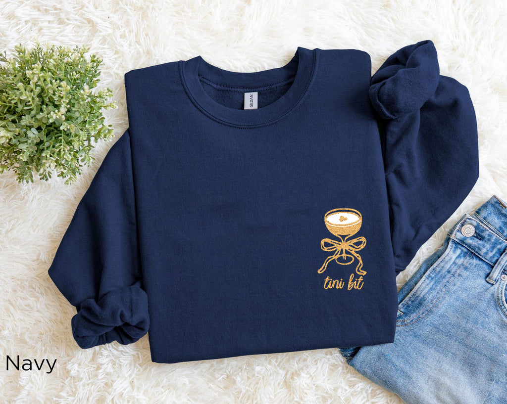 Folded navy “A Tini Bit Older” embroidered martini sweatshirt, rolled sleeves, paired with denim jeans, fun and cozy women’s birthday or party outfit.